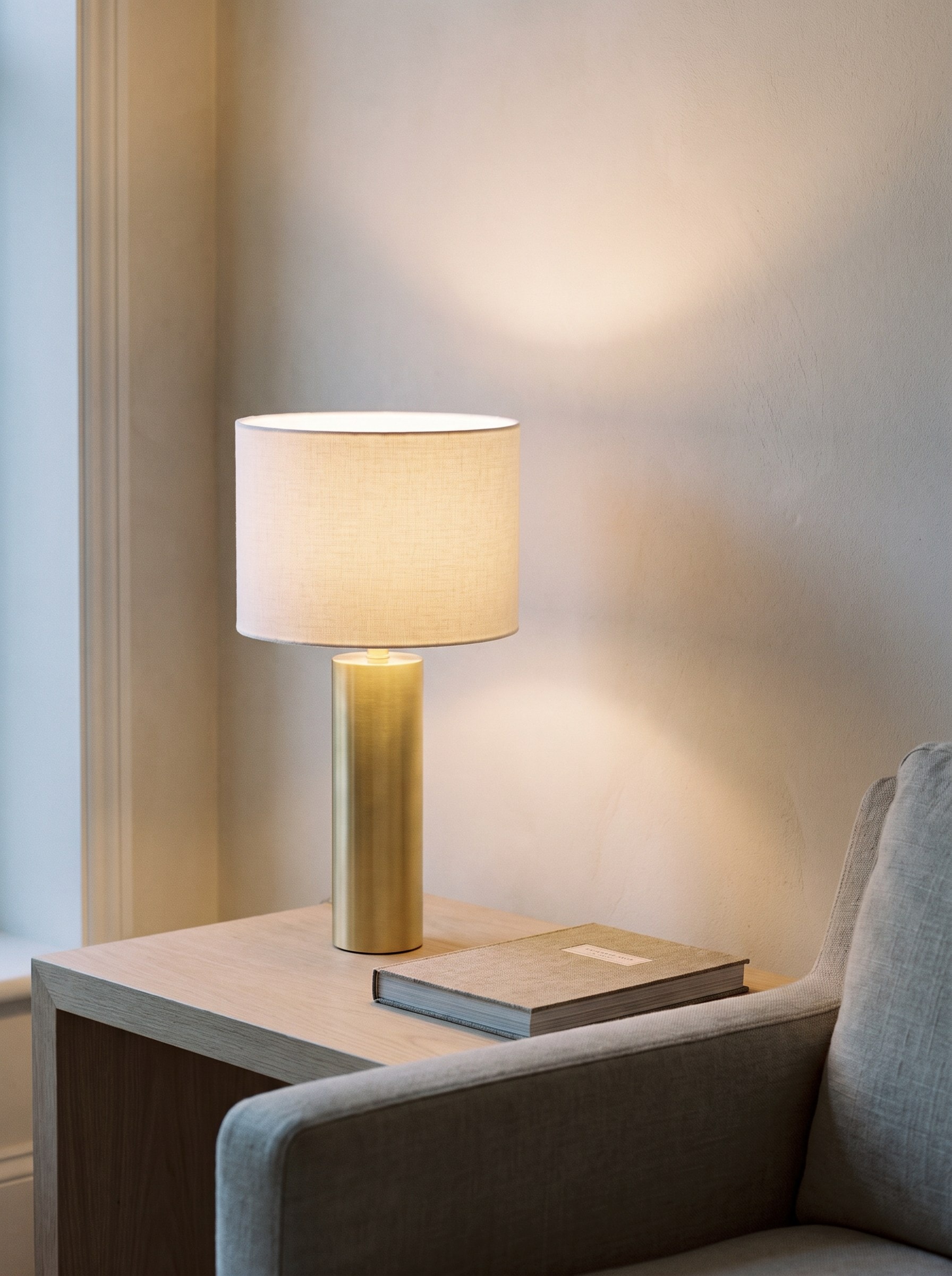 The Aalto Table Lamp lit on a pale oak side table in the corner of a quiet room, its warm light pooling on the plaster wall behind the linen shade, a book beside it and the arm of a linen chair at the edge of the frame.
