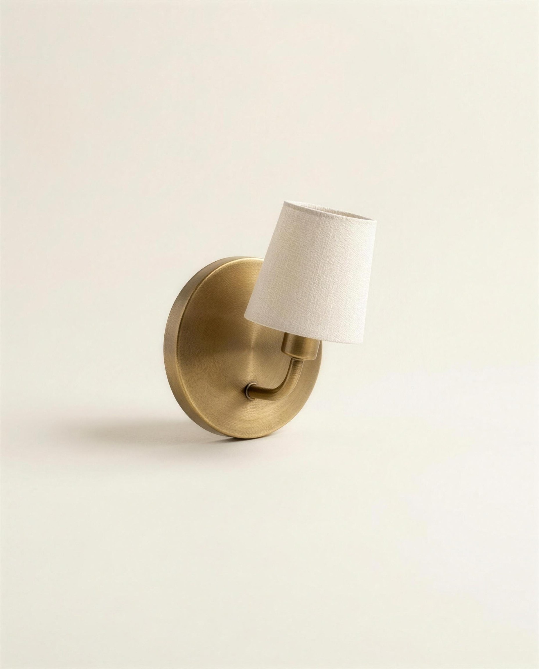 The Marlow Wall Sconce: a round solid brass backplate with a curved arm carrying a small tilted off-white linen shade.
