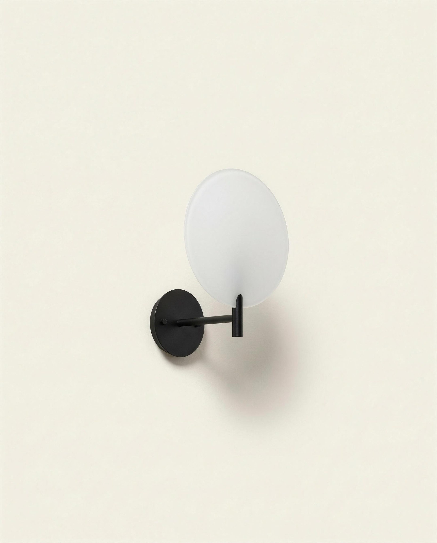The Ora Sconce: a flat opal glass disc held away from the wall on a slim blackened steel arm and round backplate.