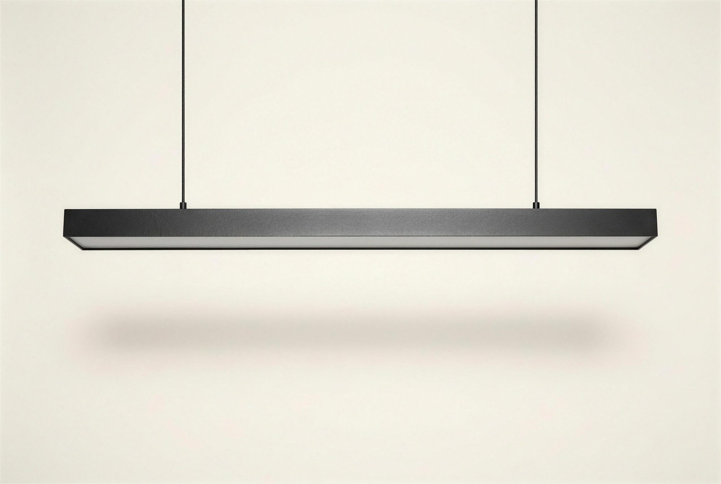 The Rill Linear Pendant: a long narrow blackened steel channel with a frosted lens, suspended horizontally on two thin cables.
