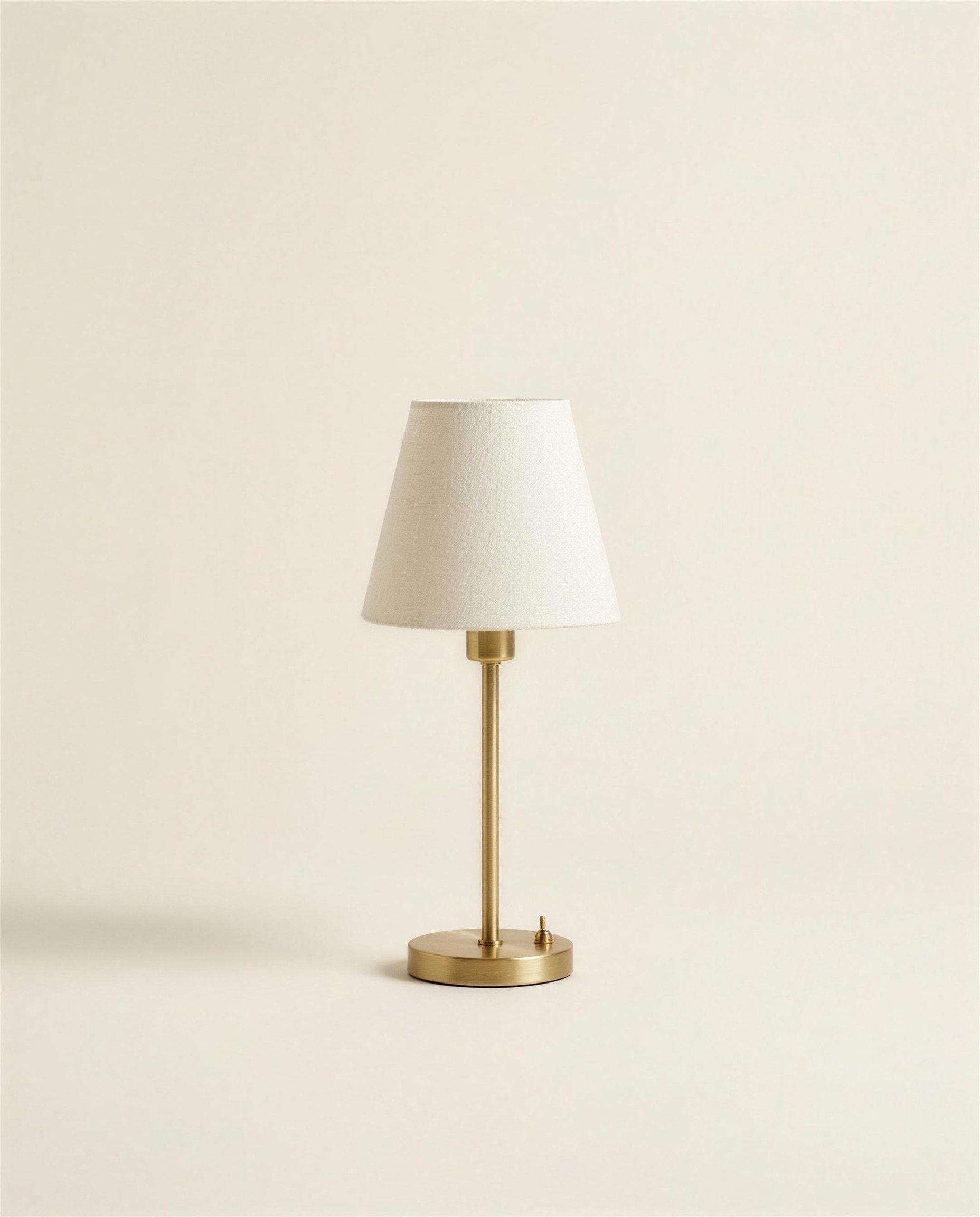 The Rowe Bedside Light: a slim brass stem on a round brass base under a tapered off-white linen shade.