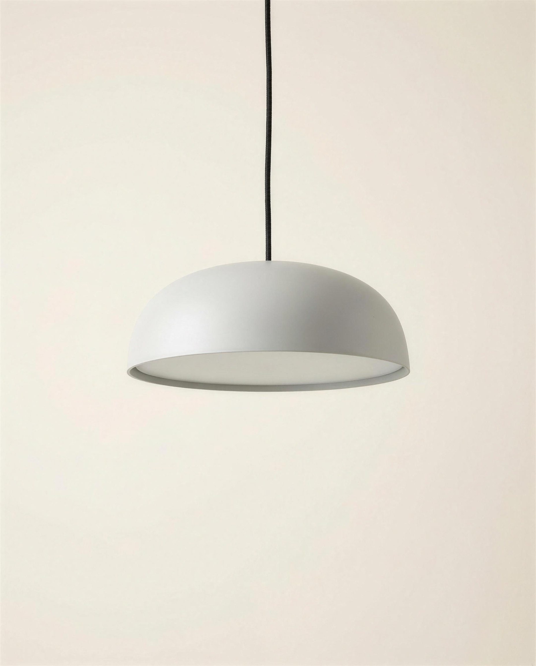 The Solen Pendant: a shallow wide spun aluminium dome in matte pale grey with a recessed opal glass diffuser on a black cloth cord.
