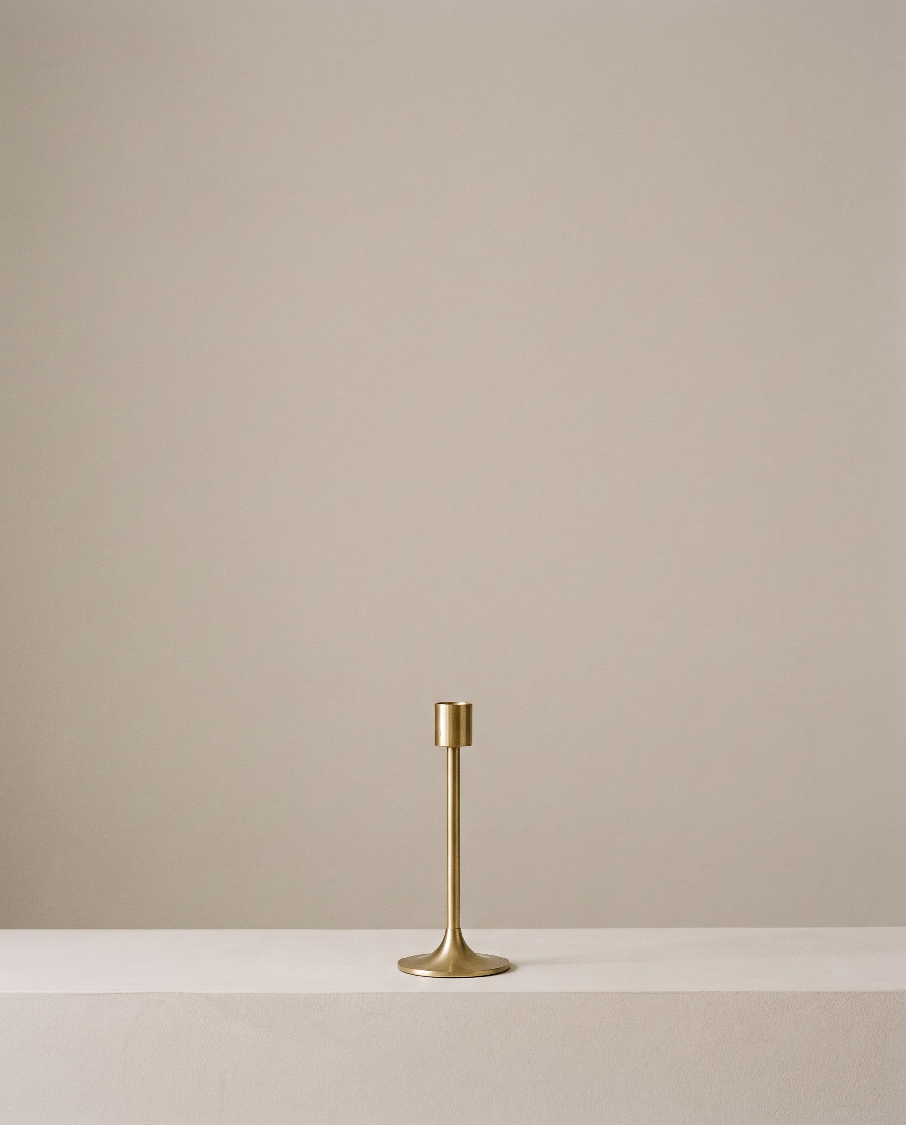 A slim brass candlestick, unlit, standing alone on a pale plaster ledge with an empty stone-grey wall rising above it.