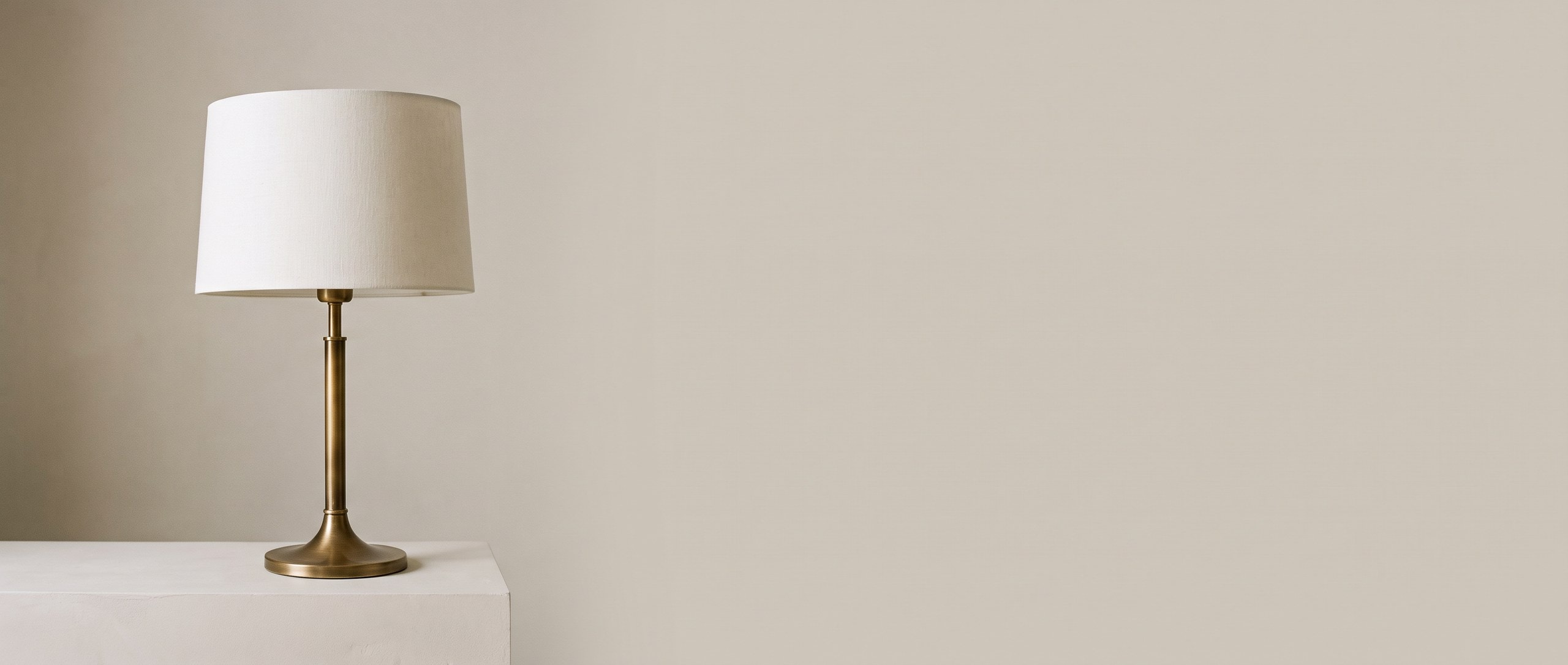A brass table lamp with an off-white linen drum shade, unlit, standing alone at the left of a plaster ledge against a plain stone-grey wall.
