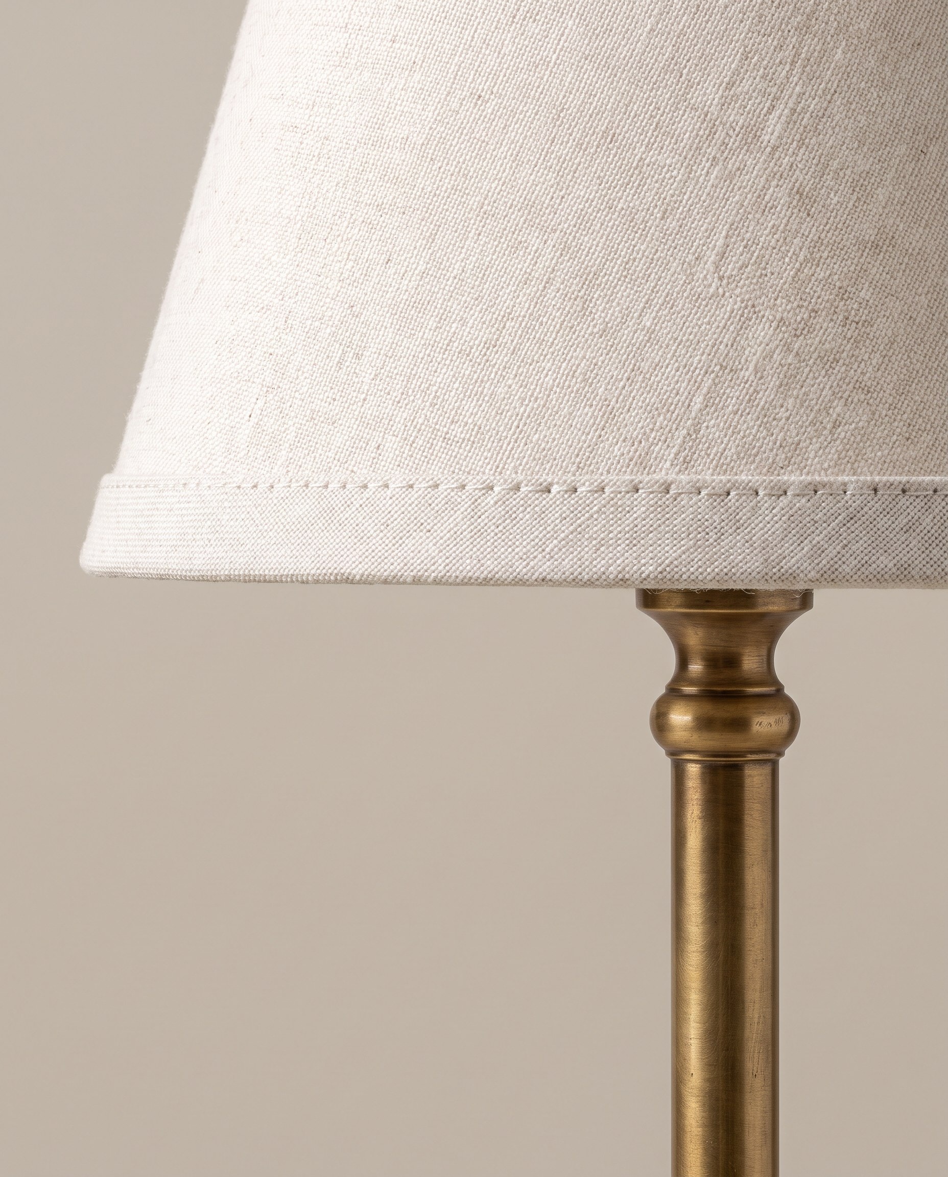 Close view of the stitched lower edge of an off-white linen lampshade meeting the turned brass stem beneath it.