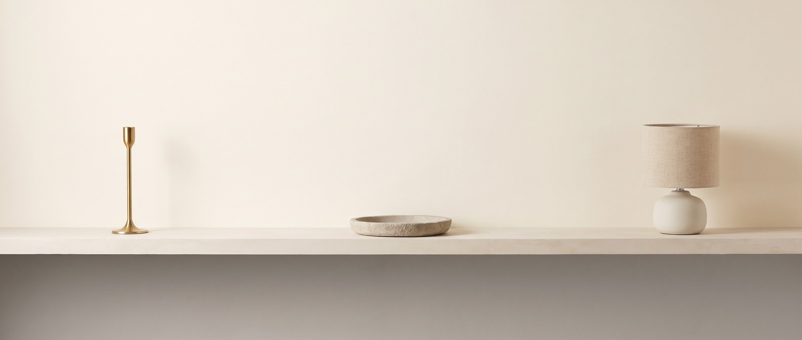 Three unlit objects spaced far apart along a pale plaster shelf: a slim brass candlestick, a shallow stone tray, and a small table light with a ceramic base and linen shade.